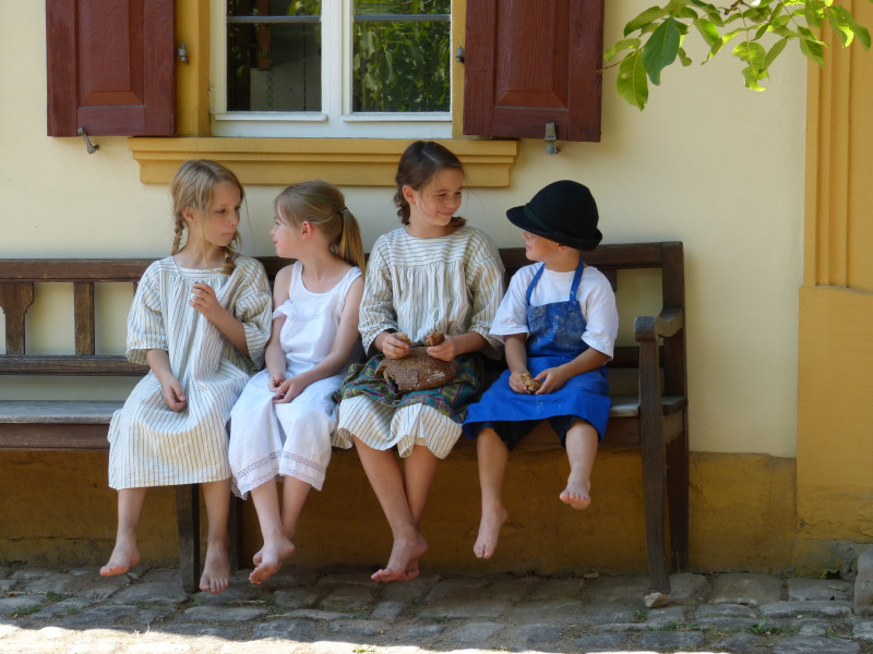 Vier Kinder in historischen Kleidern sitzen auf einer Bank vor einem Museumsgebäude.
